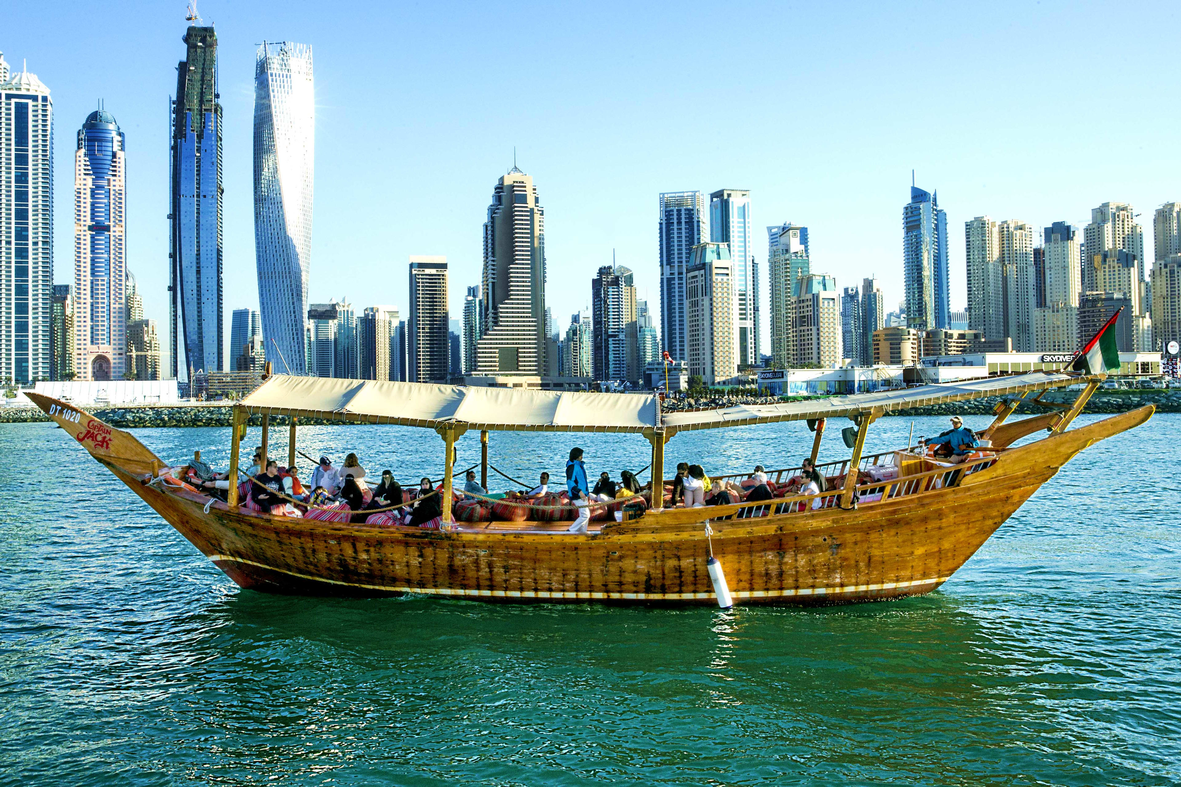 Traditional wooden dhow boat with passengers sailing on turquoise water with Dubai skyline skyscrapers in background